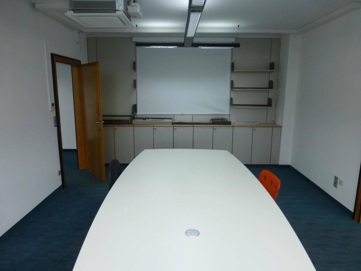 Konferenzraum mit großem Tisch, integrierten Schränken und Projektionsfläche, ideal für Präsentationen.