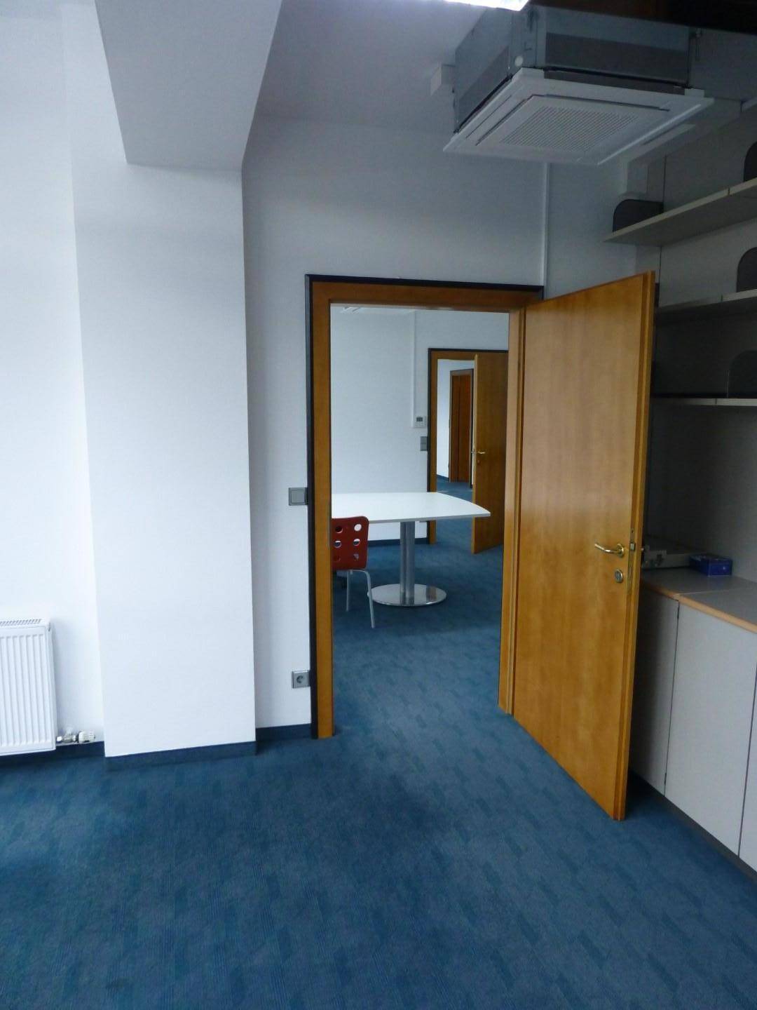 Blick durch eine offene Holztür in einen weiteren Bürobereich mit Tisch und Teppichboden.