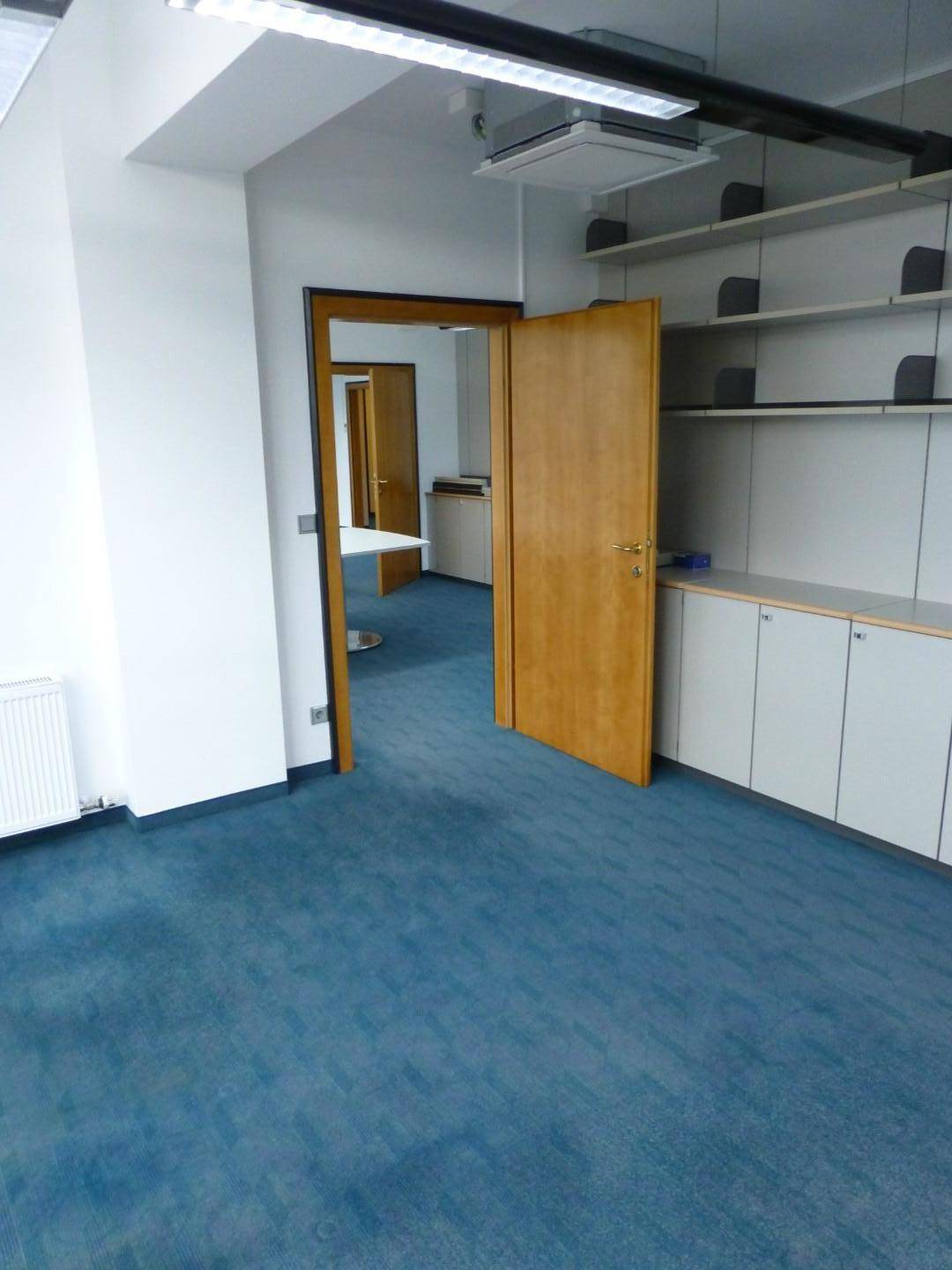 Offene Holztür führt in einen Bürobereich mit Einbauregalen und blauem Teppichboden.