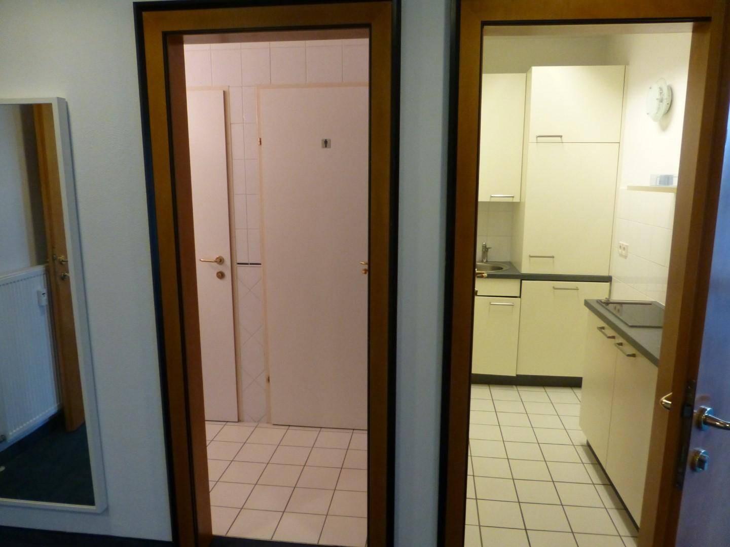 Blick durch zwei offene Türen in einen gefliesten Sanitärbereich und eine kleine Küchenzeile.
