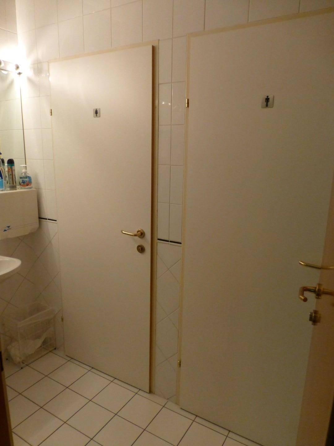 Sanitärbereich mit zwei Toilettenkabinen, Waschbecken und weißen Fliesen an Wänden und Boden.