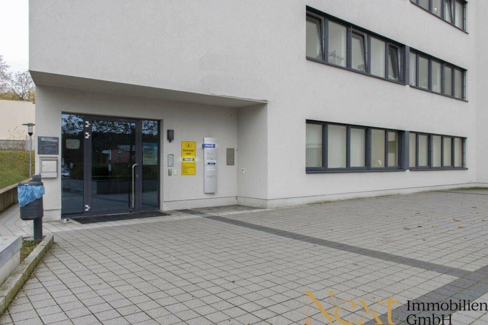 Eingangsbereich eines modernen Bürogebäudes mit Glasfassade und gepflastertem Vorplatz.