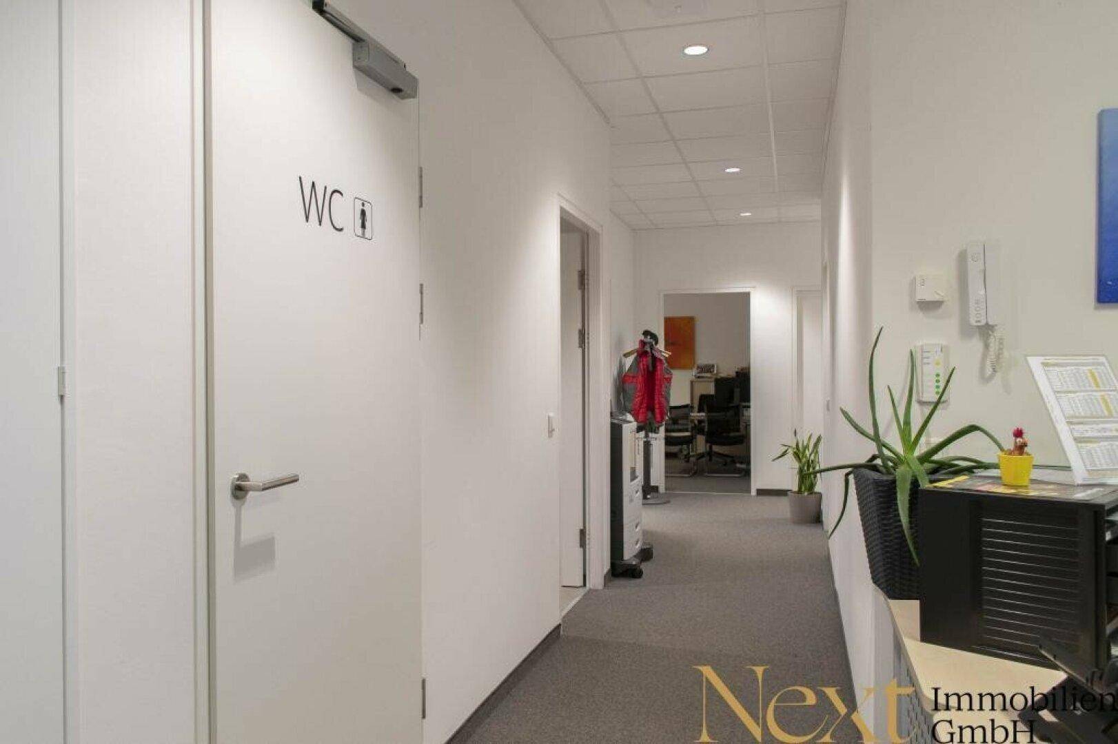 Heller Flur mit Türen, darunter eine Toilette, und einem Blick in einen weiteren Bürobereich.