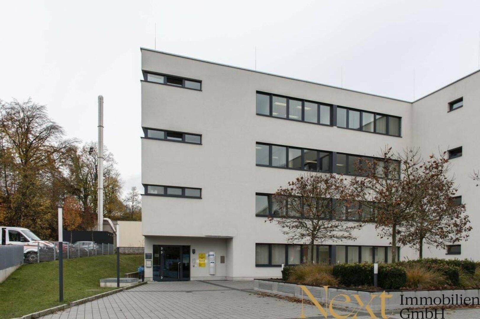 Modernes Bürogebäude mit weißer Fassade, vielen Fenstern und gepflegter Außenanlage mit Bäumen.