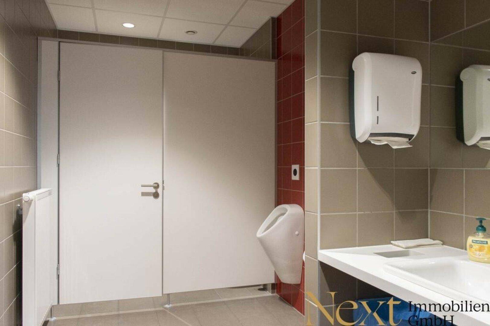 Öffentliche Toilette mit Urinalen, Toilettenkabinen, Waschbecken und gefliesten Wänden in Braun- und Rottönen.
