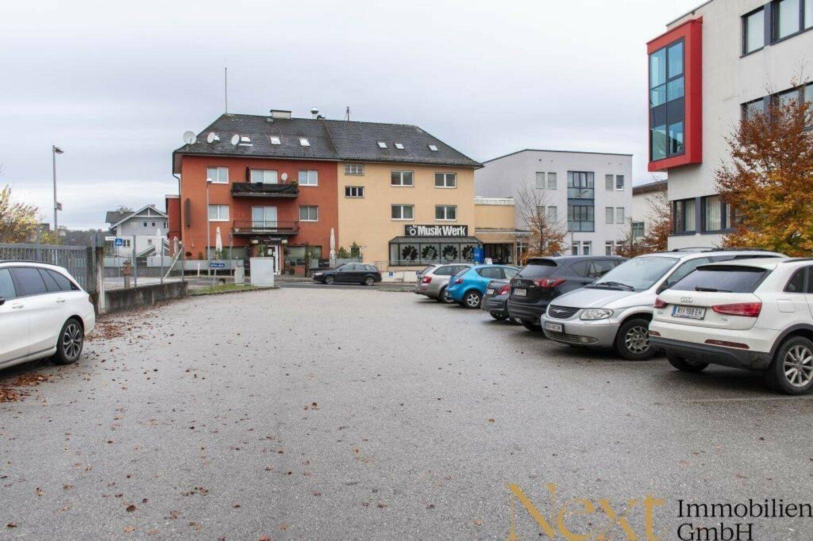 Außenansicht eines Parkplatzes mit geparkten Autos und umliegenden Gebäuden im Hintergrund.