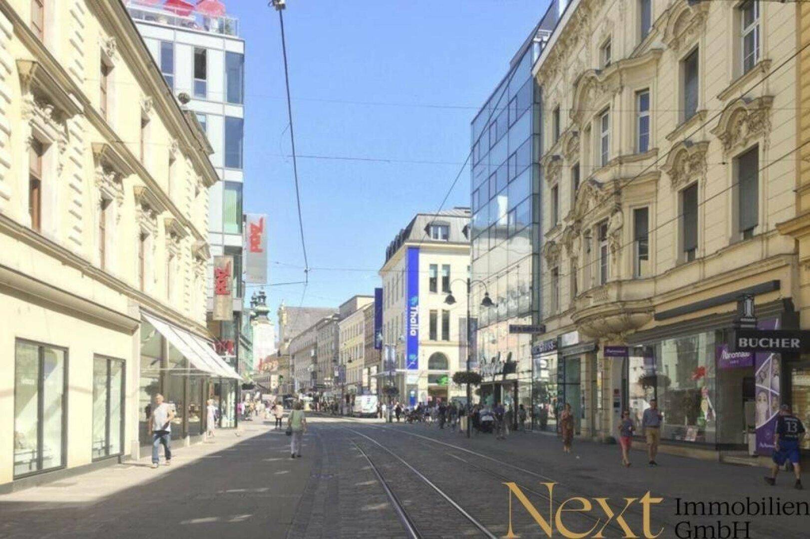 Belebte Einkaufsstraße mit historischen Gebäuden, modernen Fassaden und Straßenbahnschienen unter blauem Himmel.
