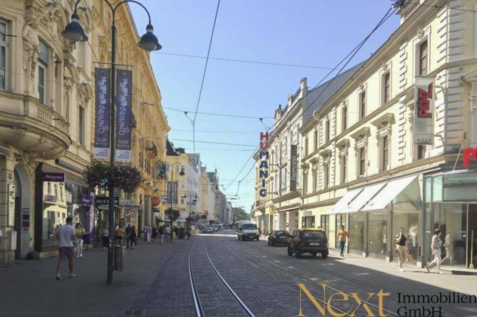 Belebte Stadtstraße mit historischen Gebäuden, Geschäften und Straßenbahnschienen unter klarem Himmel.