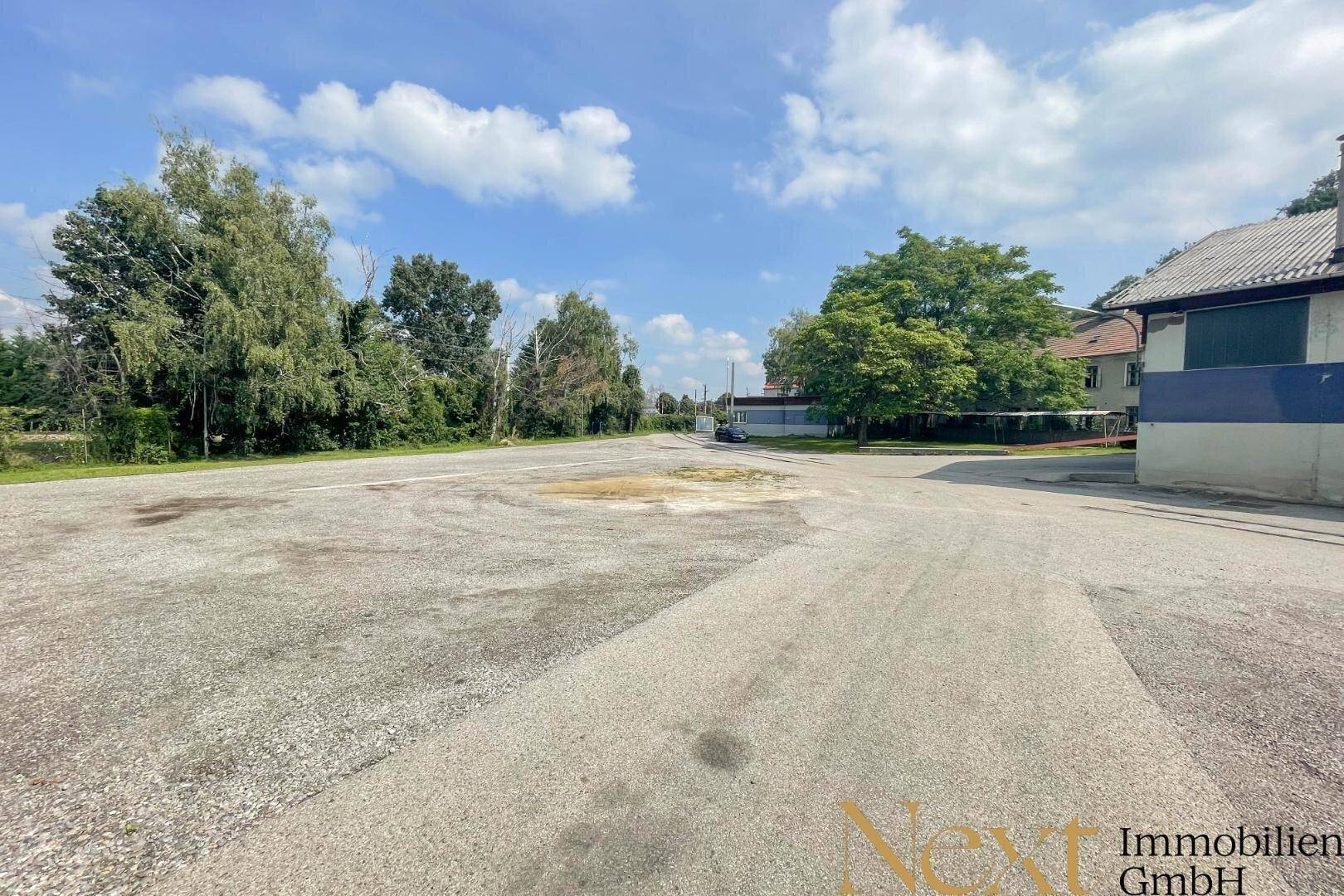 Großer Schotterparkplatz unter blauem Himmel mit vereinzelten Bäumen und Gebäuden im Hintergrund.