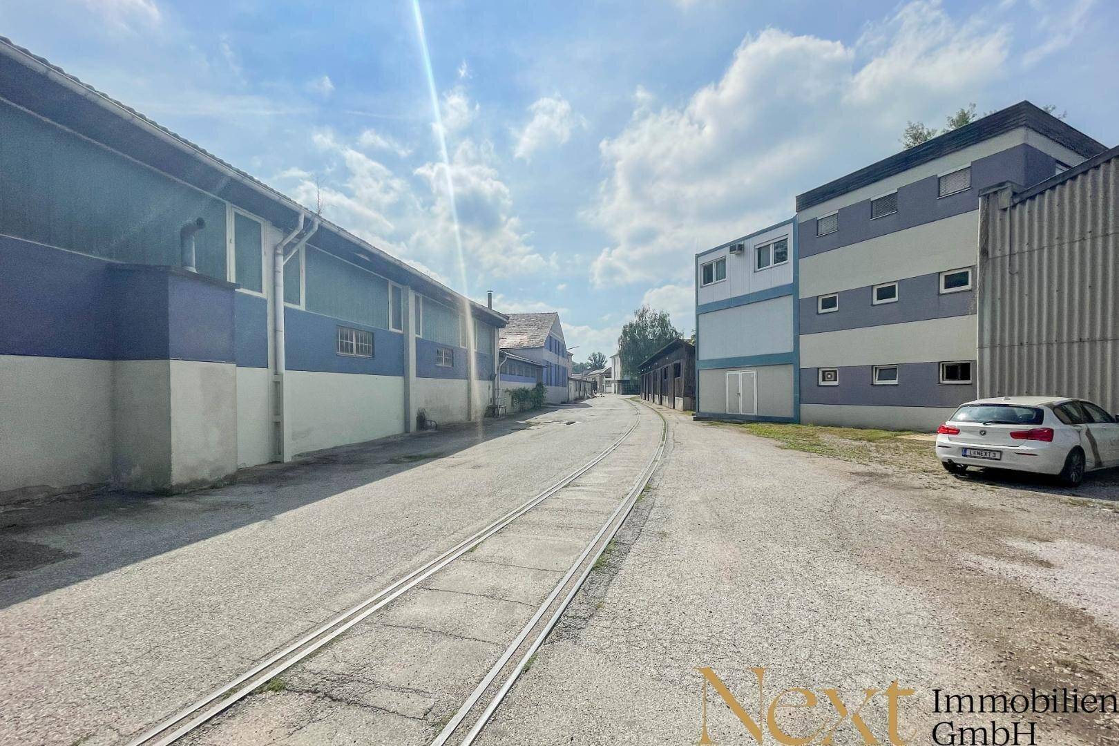 Zufahrtsstraße mit alten Bahnschienen, umgeben von Industriegebäuden unter einem bewölkten Himmel.