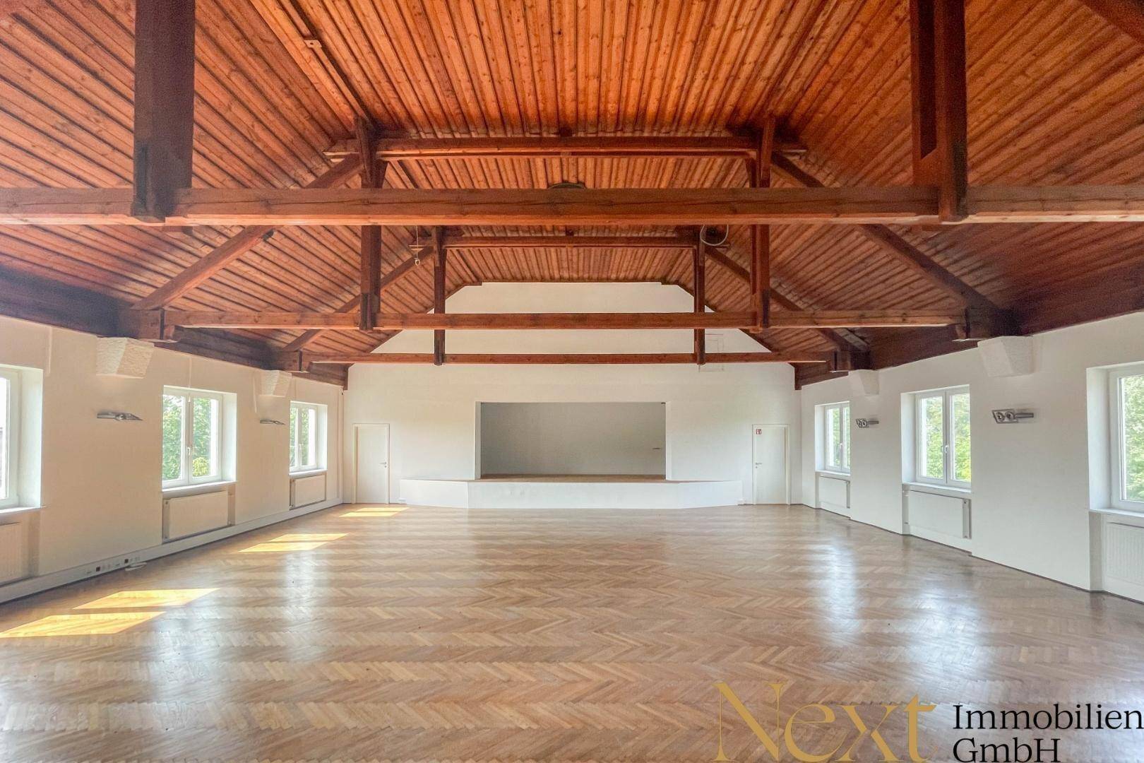 Großer Saal mit Holzbalkendecke und Fischgrätparkett, ideal für Veranstaltungen und Versammlungen.