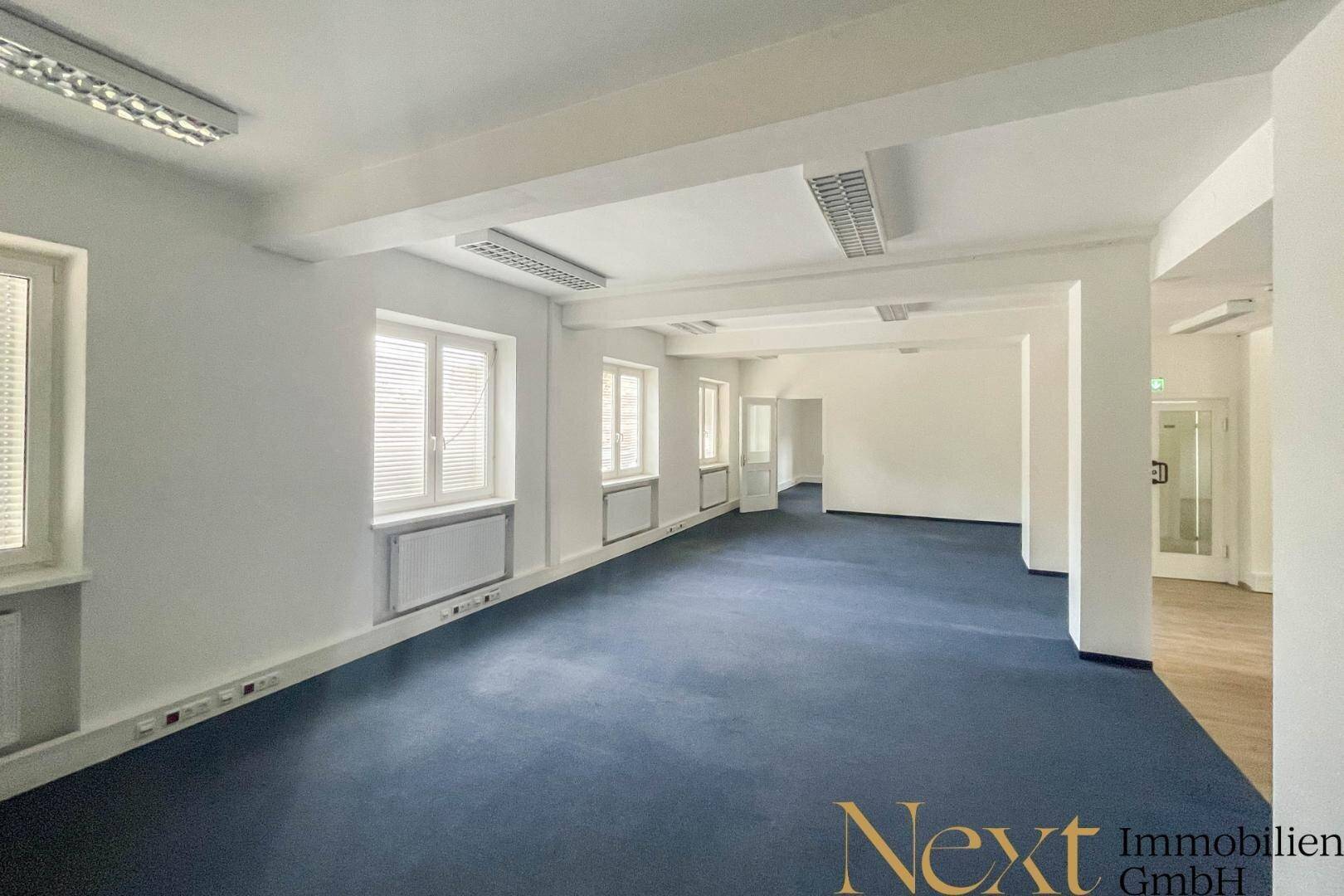 Großes, offenes Büro mit blauem Teppichboden und vielen Fenstern, geeignet für mehrere Arbeitsplätze.