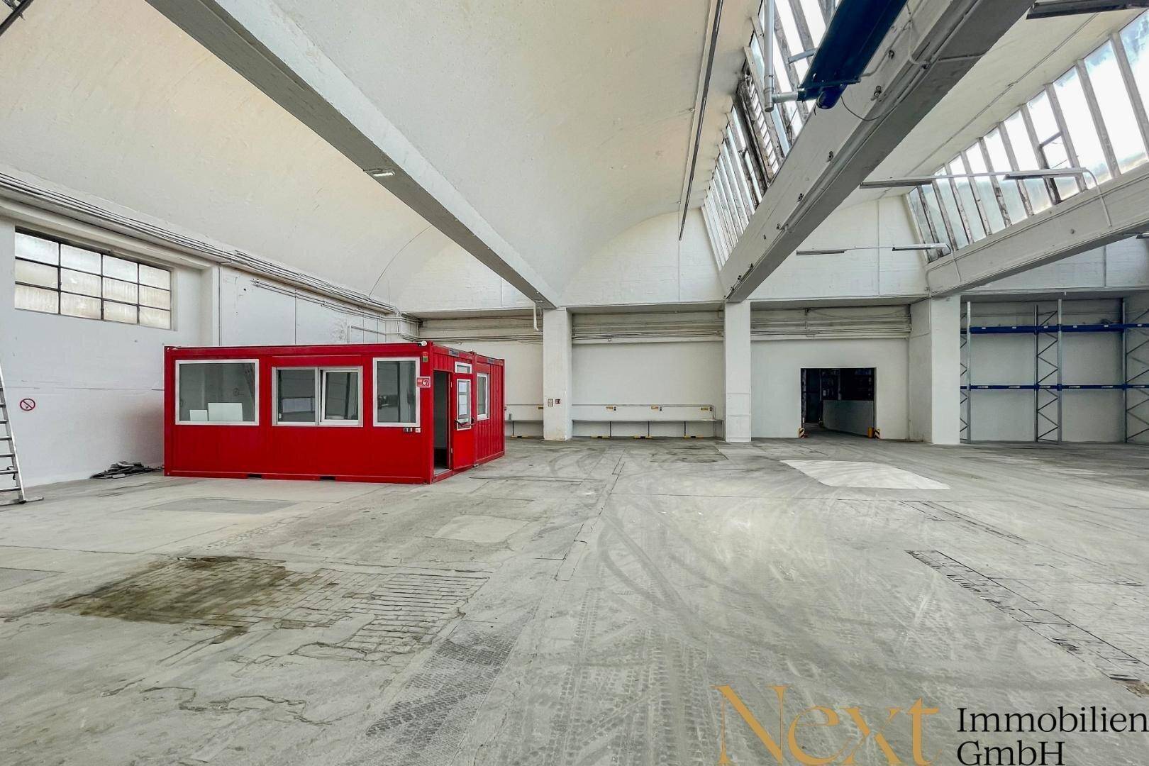 Weitläufige Lagerhalle mit Betonboden und einem roten Bürocontainer, gut beleuchtet durch Oberlichter.