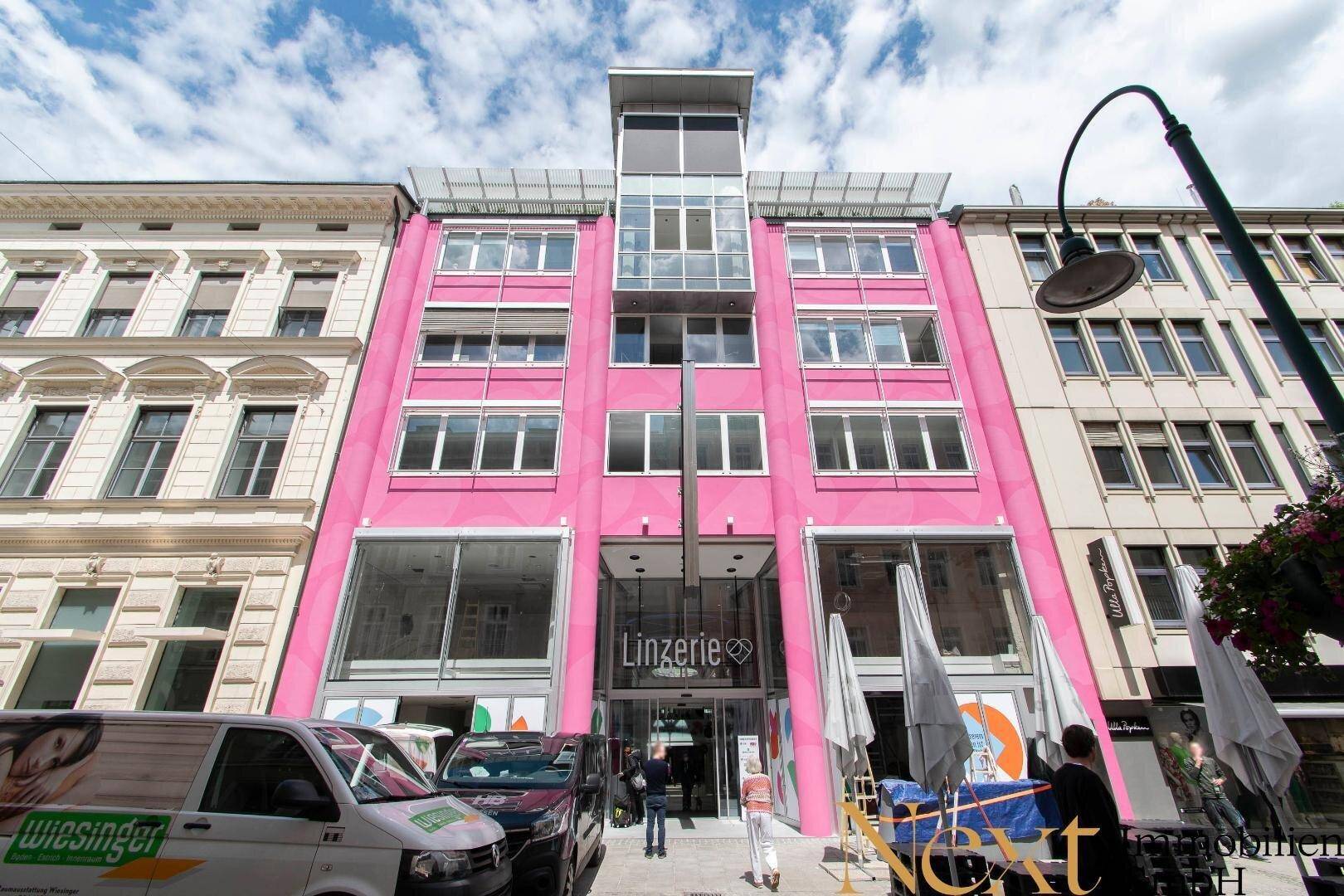 Modernes Gebäude mit auffälliger pinker Fassade, großen Fenstern und Einzelhandelsflächen im Erdgeschoss.