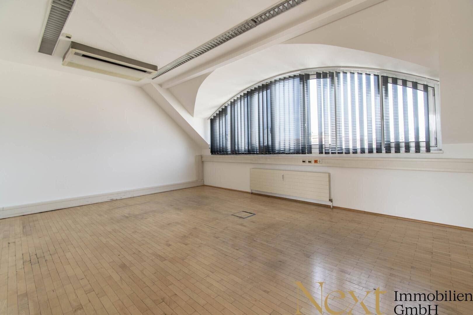 Heller Büroraum im Dachgeschoss mit gebogenem Fenster, Jalousien und glänzendem Holzboden.