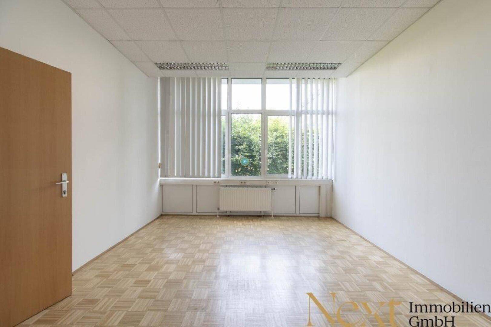 Helles Büro mit Parkettboden und großen Fenstern, die mit vertikalen Jalousien ausgestattet sind.