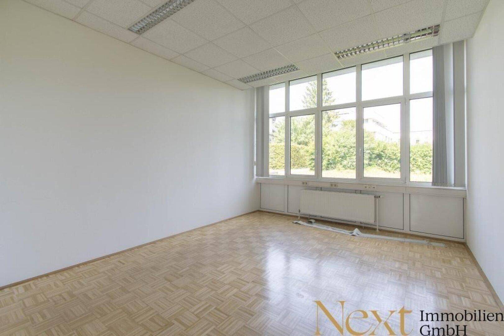 Geräumiges Büro mit Parkettboden und großen Fenstern, die einen grünen Ausblick bieten.