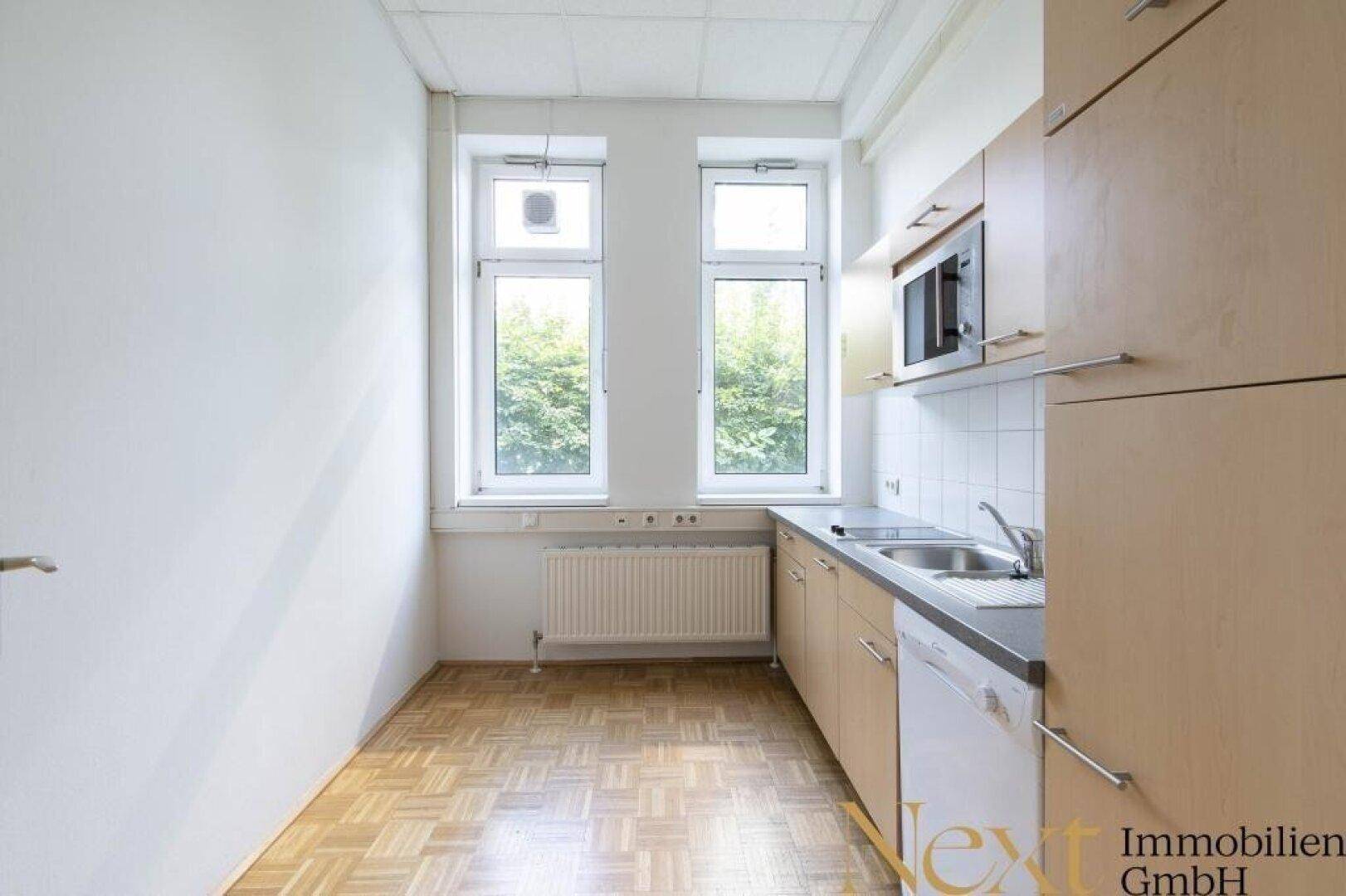Kleine Küchenzeile mit Spüle, Mikrowelle und Parkettboden in einem hellen Raum mit Fenstern.