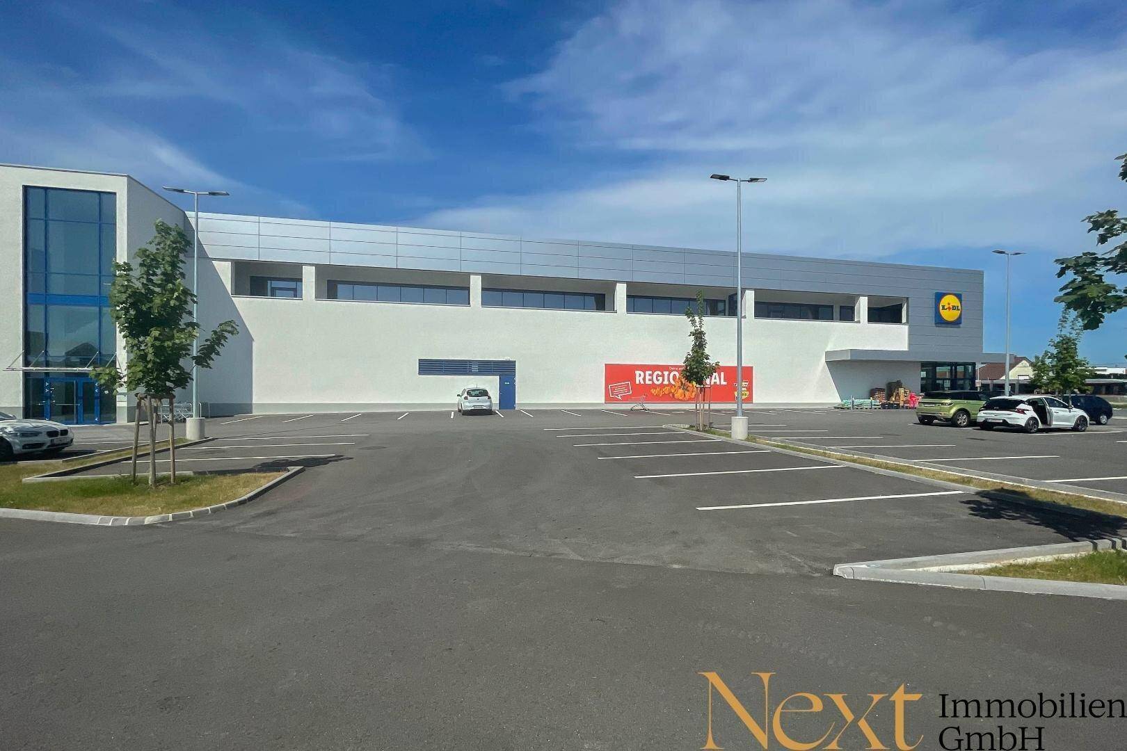 Weitläufiger Blick auf die moderne Supermarkt-Fassade und den großzügigen Parkplatz an einem sonnigen Tag.