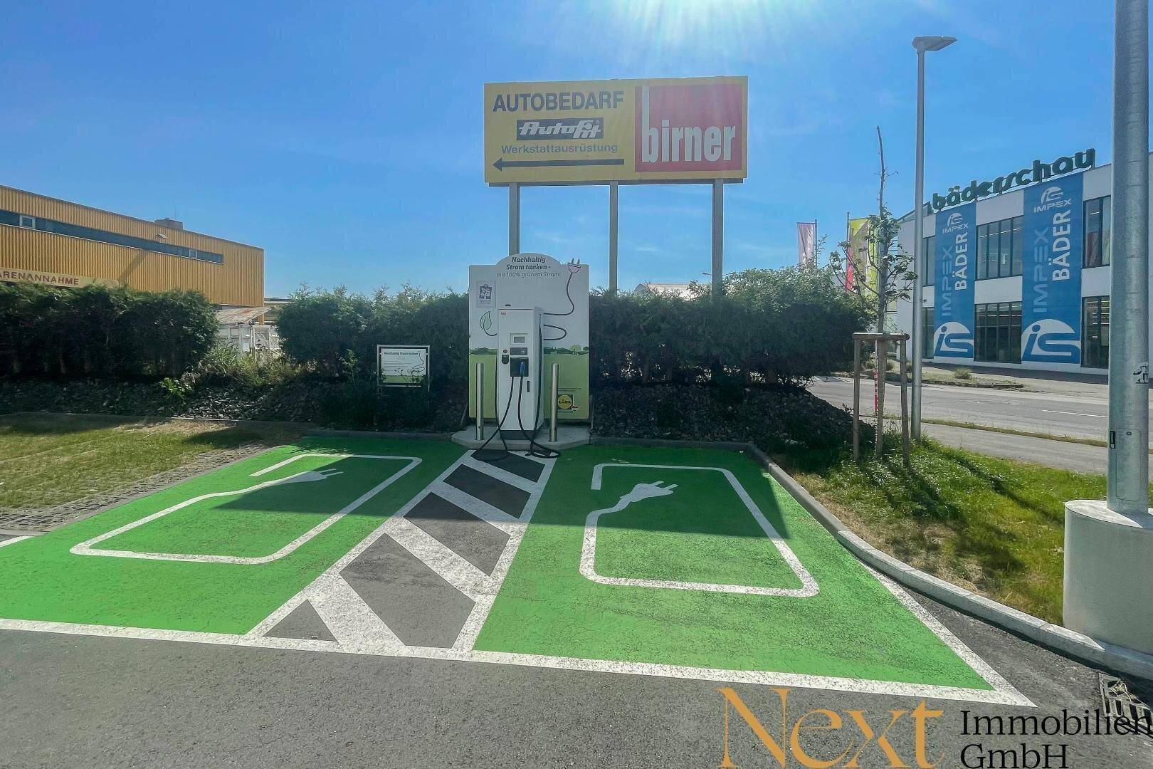 Moderne Ladestation für Elektrofahrzeuge auf einem Parkplatz mit grüner Markierung und Werbeschildern im Hintergrund.