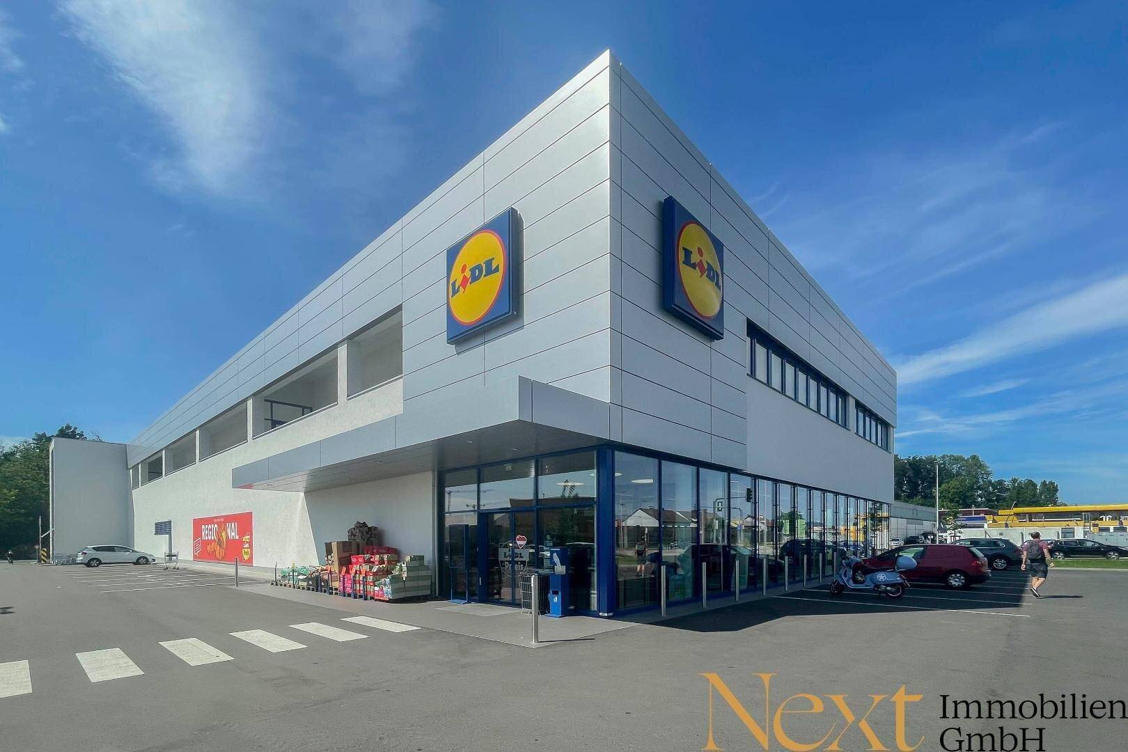 Eckansicht des modernen Supermarkts mit großflächigen Fenstern, Haupteingang und angrenzendem Parkplatz.