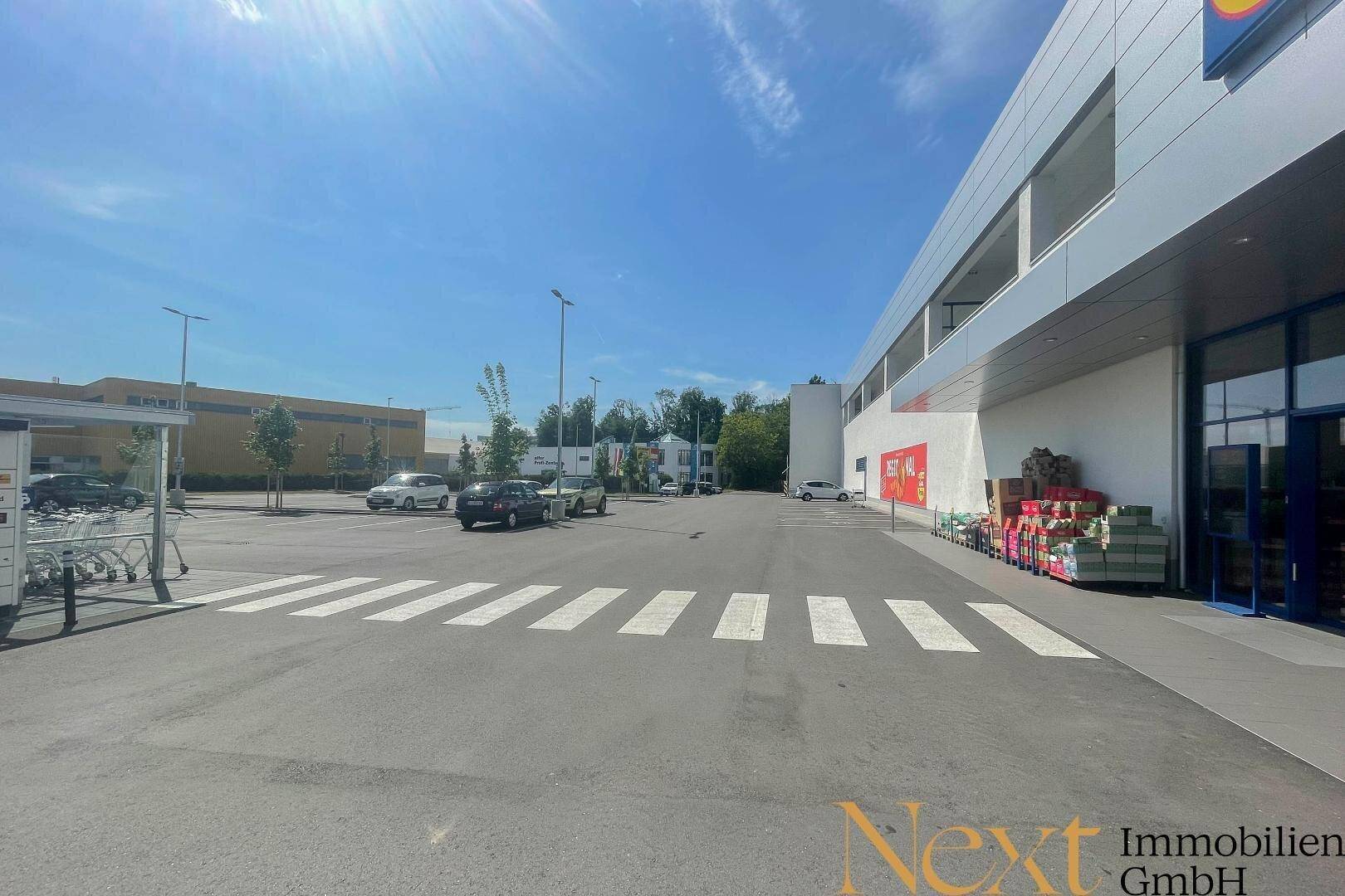 Blick über den Parkplatz mit Zebrastreifen zur Seite des modernen Supermarktgebäudes unter blauem Himmel.
