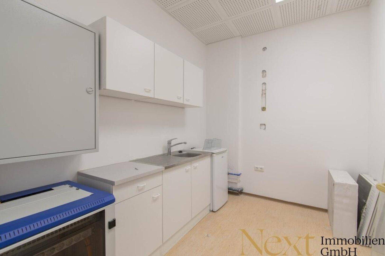 Kleine Küchenzeile mit weißen Schränken, Spüle und Kühlschrank, ideal für Pausenbereiche in einem Büro.