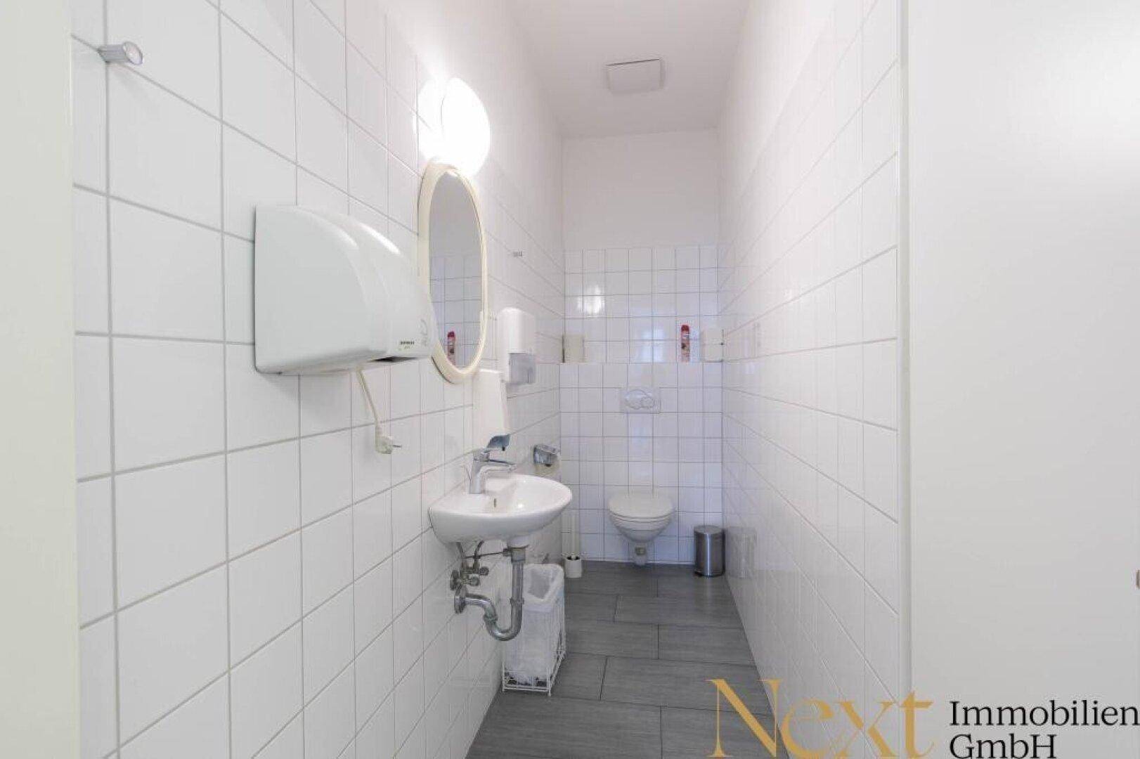 Saubere, geflieste Toilette mit Waschbecken, WC und Handtuchspender, in gutem und funktionalem Zustand.