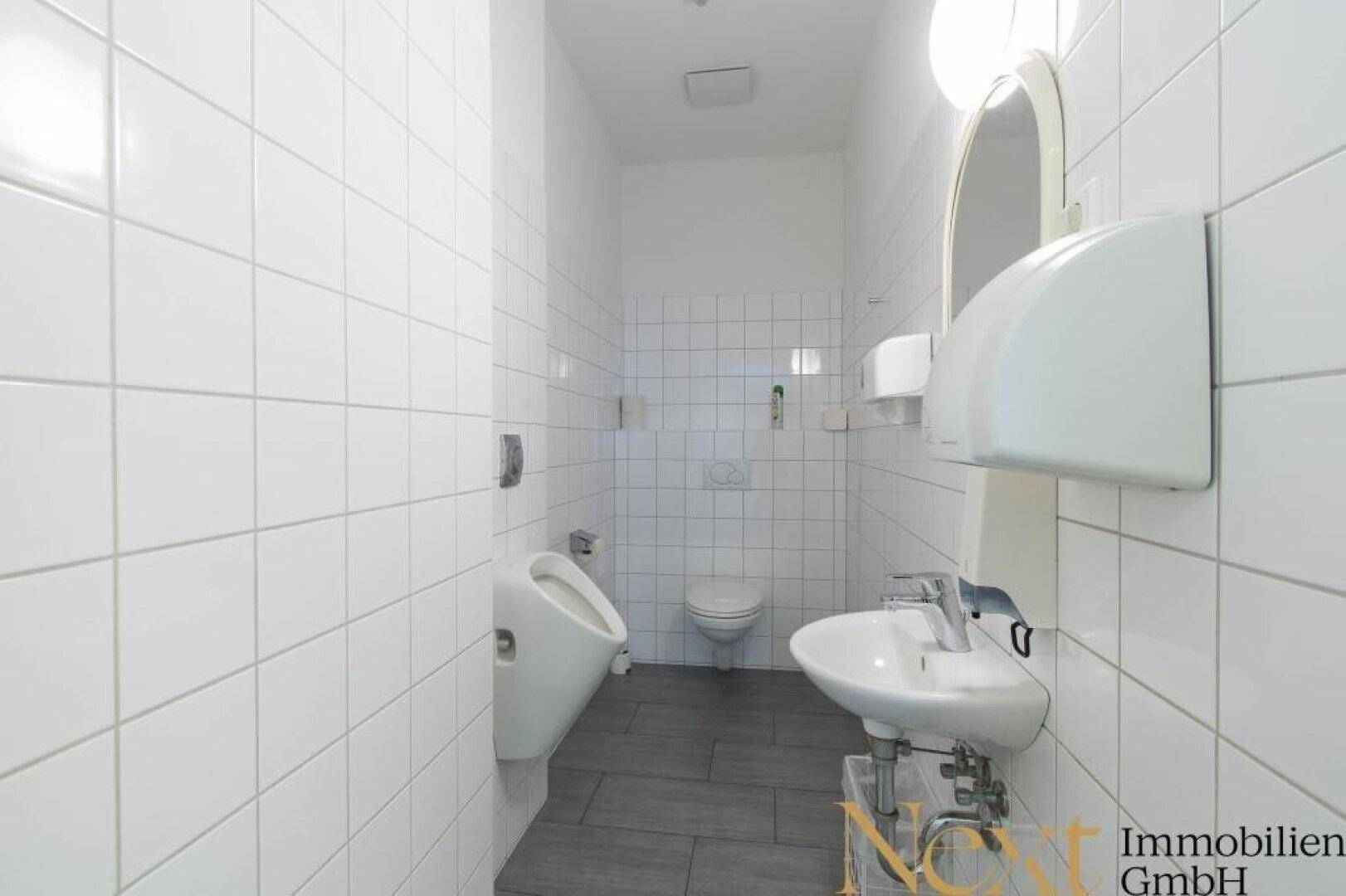 Geflieste Herrentoilette mit Waschbecken, Urinal und WC, ausgestattet mit einem Handtuchspender.