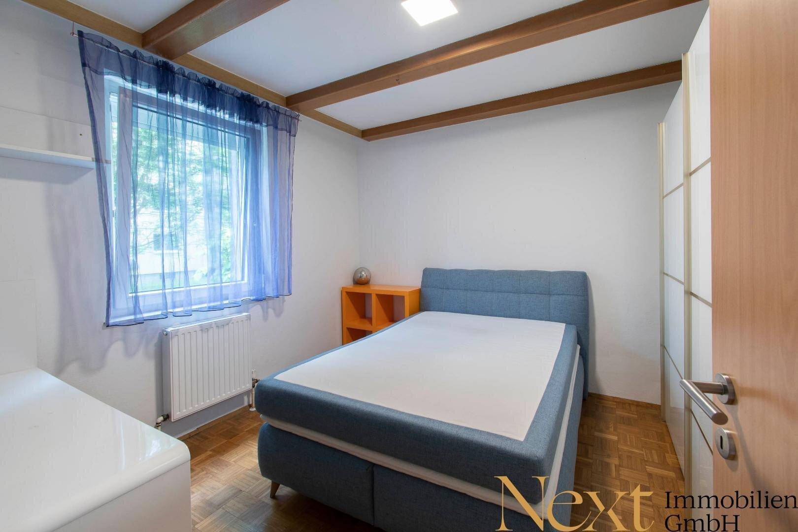 Schlafzimmer mit Holzbalken an der Decke, blauem Bett und Fenster mit Vorhang.