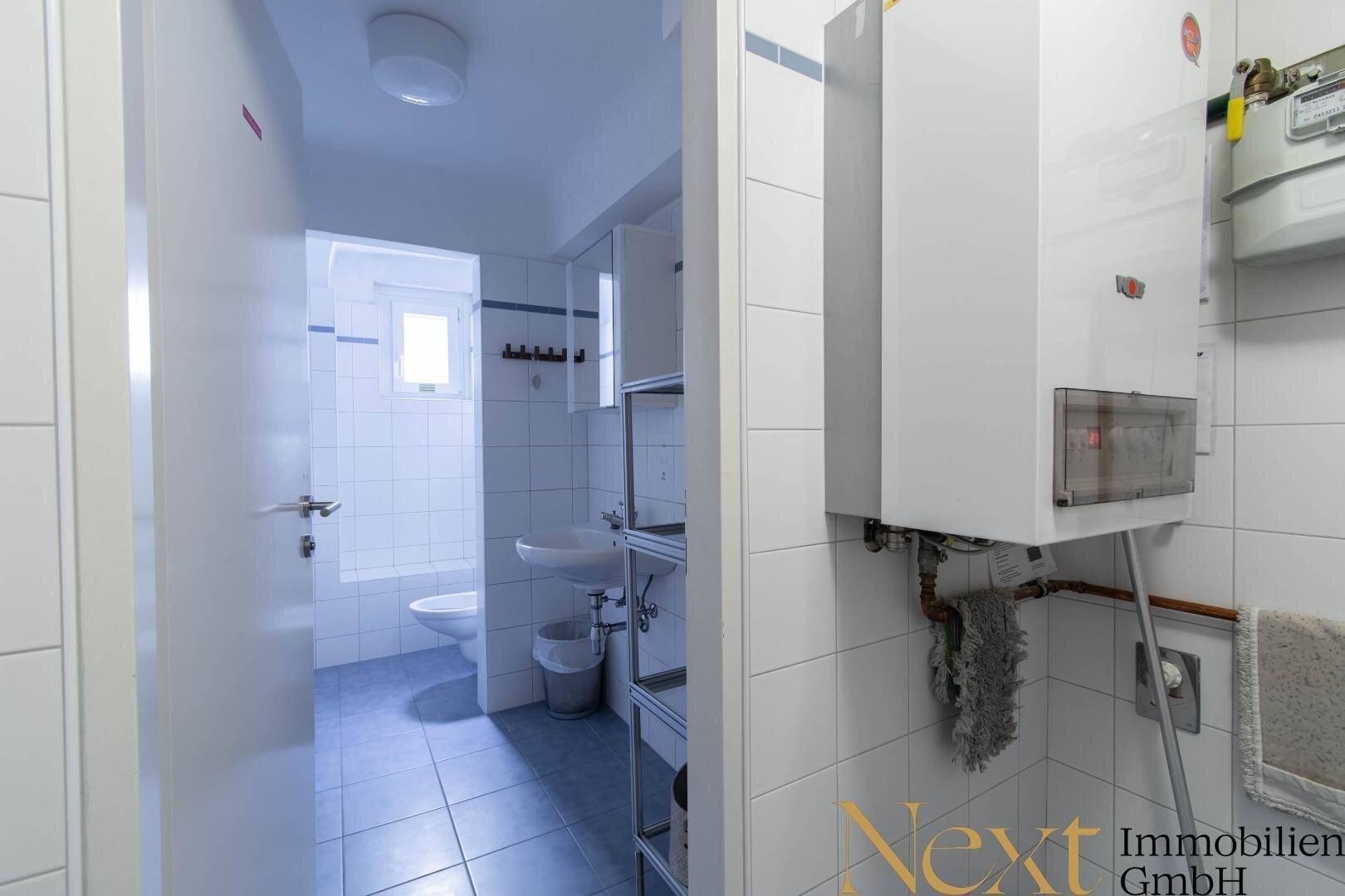 Helles Badezimmer mit weißen Fliesen, Fenster und Boiler, bietet grundlegende sanitäre Einrichtungen.