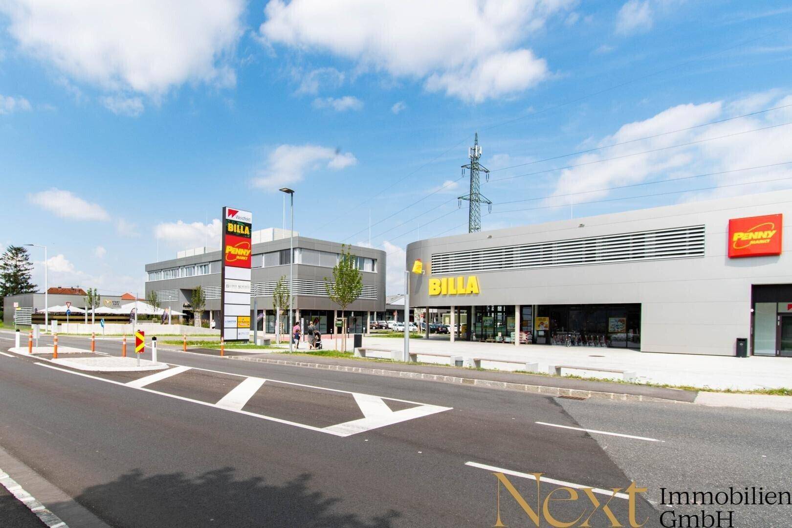Gesamtansicht eines modernen Einkaufszentrums mit Supermärkten und breiter Zufahrtsstraße.