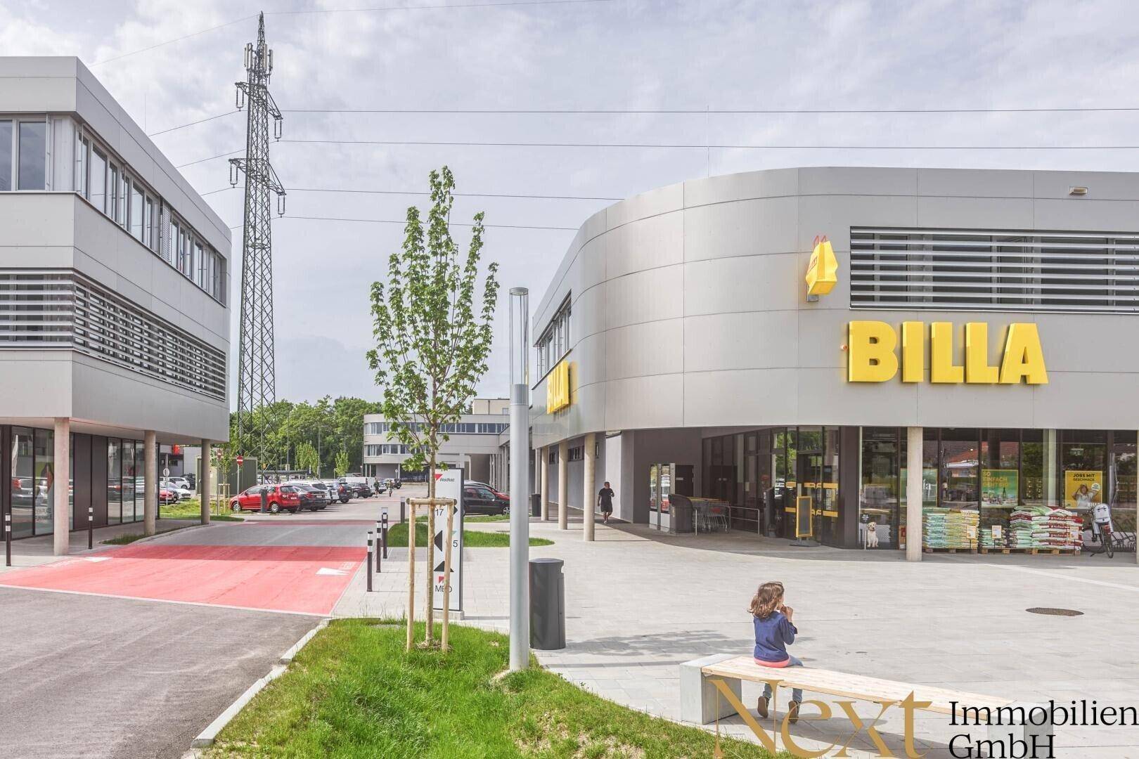Modernes Einkaufszentrum mit Supermarkt und Bürogebäude, umgeben von gepflegten Außenanlagen.