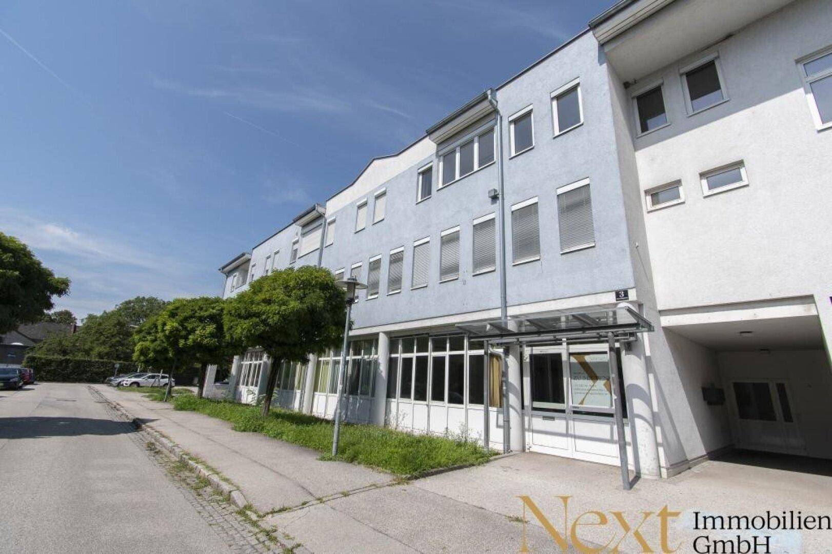 Moderne Gebäudefassade mit mehreren Etagen, großen Fenstern und Parkplätzen entlang der Straße.