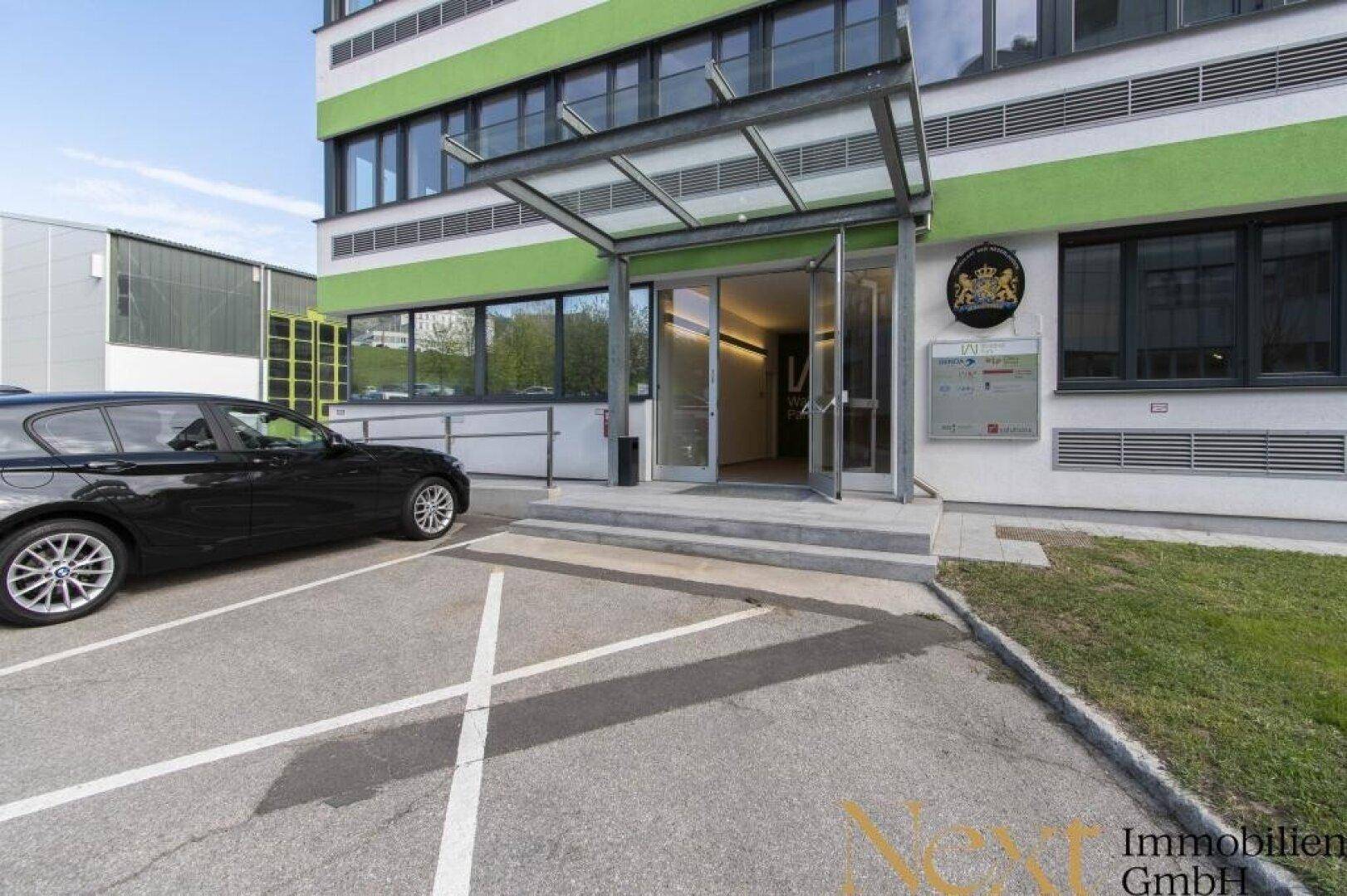 Moderner Gebäudeeingang mit Glasvordach und angrenzenden Parkplätzen, umgeben von Grünflächen.