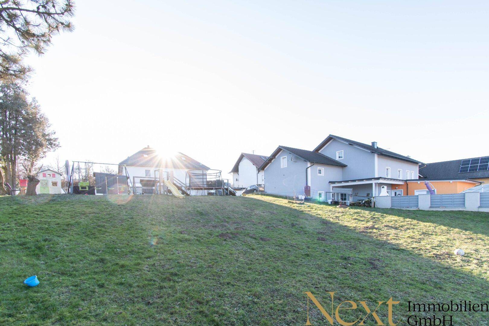 Weitläufiger Gartenbereich mit Rasenfläche und Spielgeräten im Hintergrund, unter strahlendem Sonnenschein.