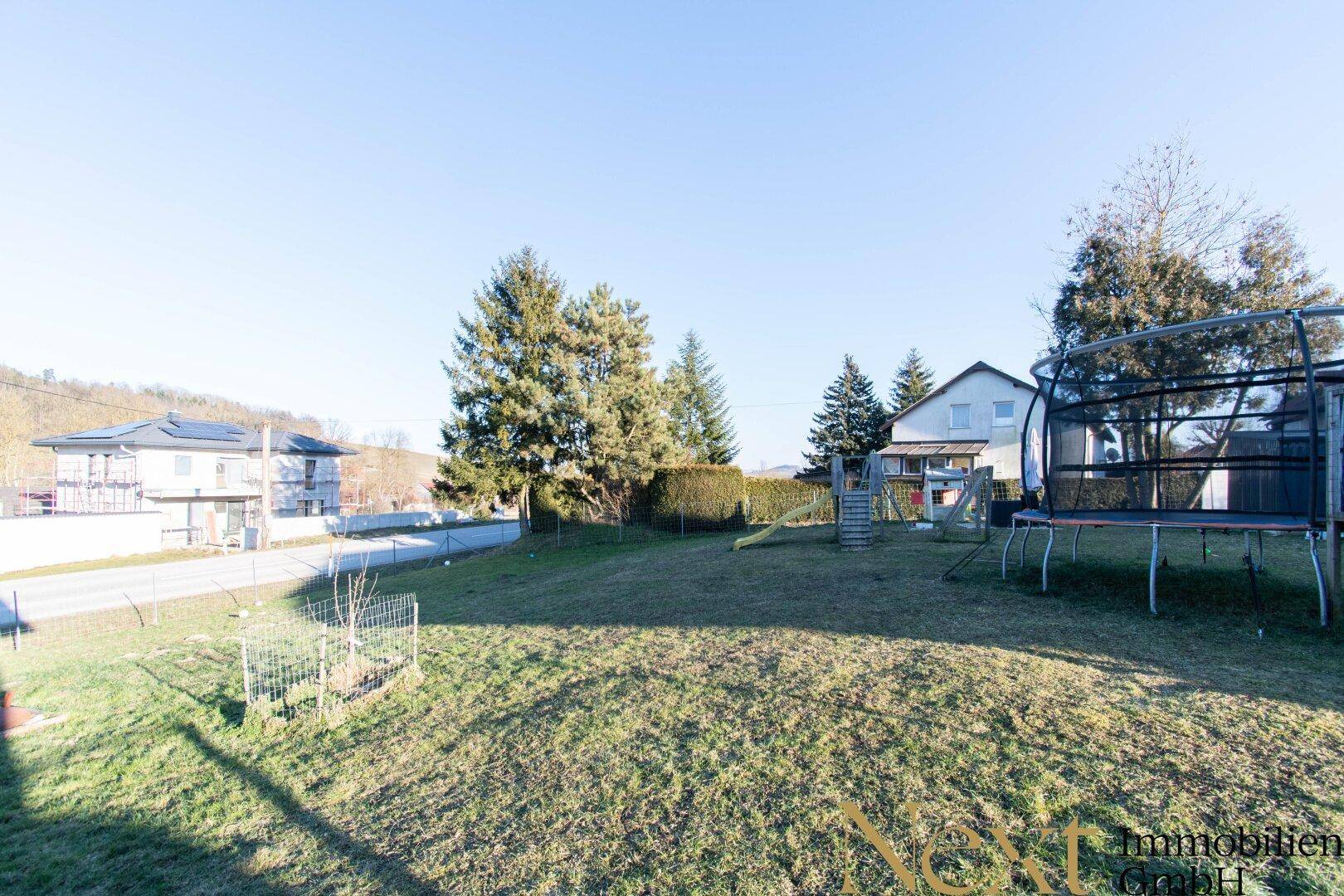 Sonniger Garten mit weitläufiger Rasenfläche, umgeben von Bäumen und mit Spielgeräten.