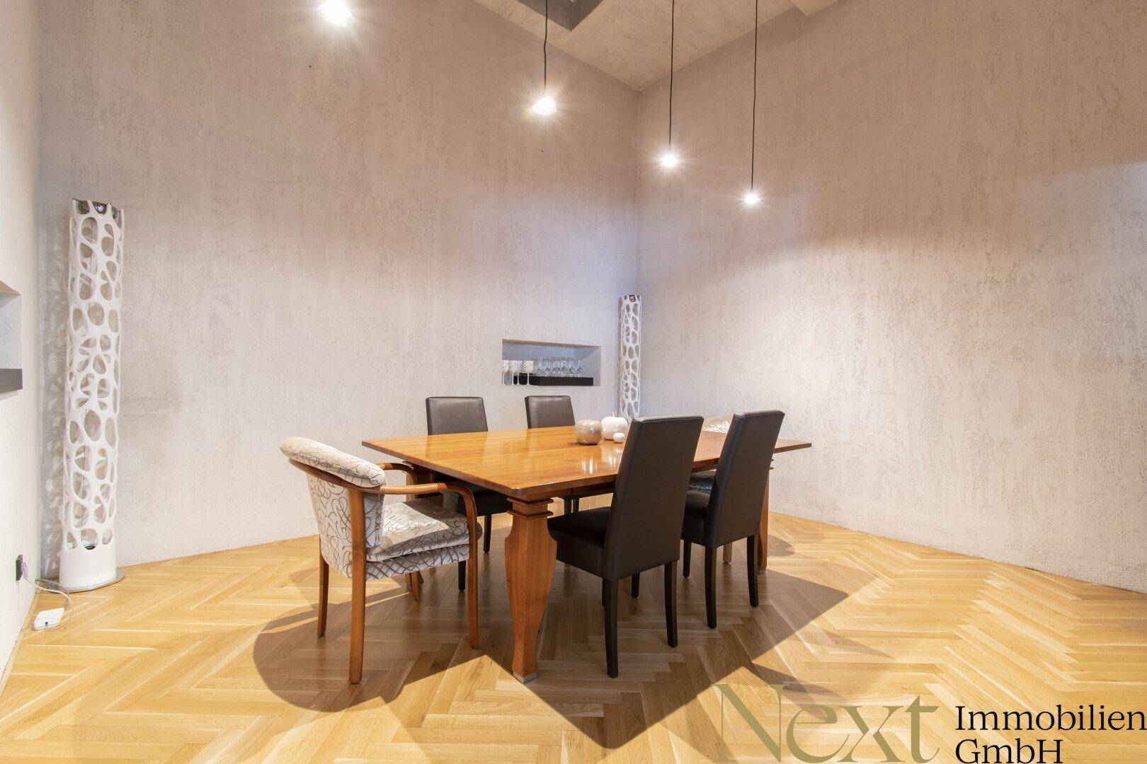 Esszimmer mit Holztisch, modernen Stühlen und Fischgrätparkett, beleuchtet von Hängelampen.
