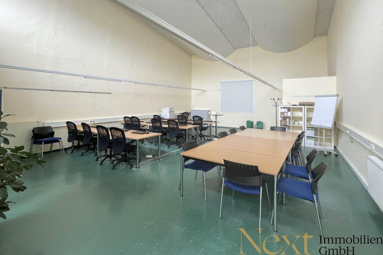 Großer Schulungsraum mit zahlreichen Tischen und Stühlen, Leinwand und Whiteboard, bereit für Präsentationen.