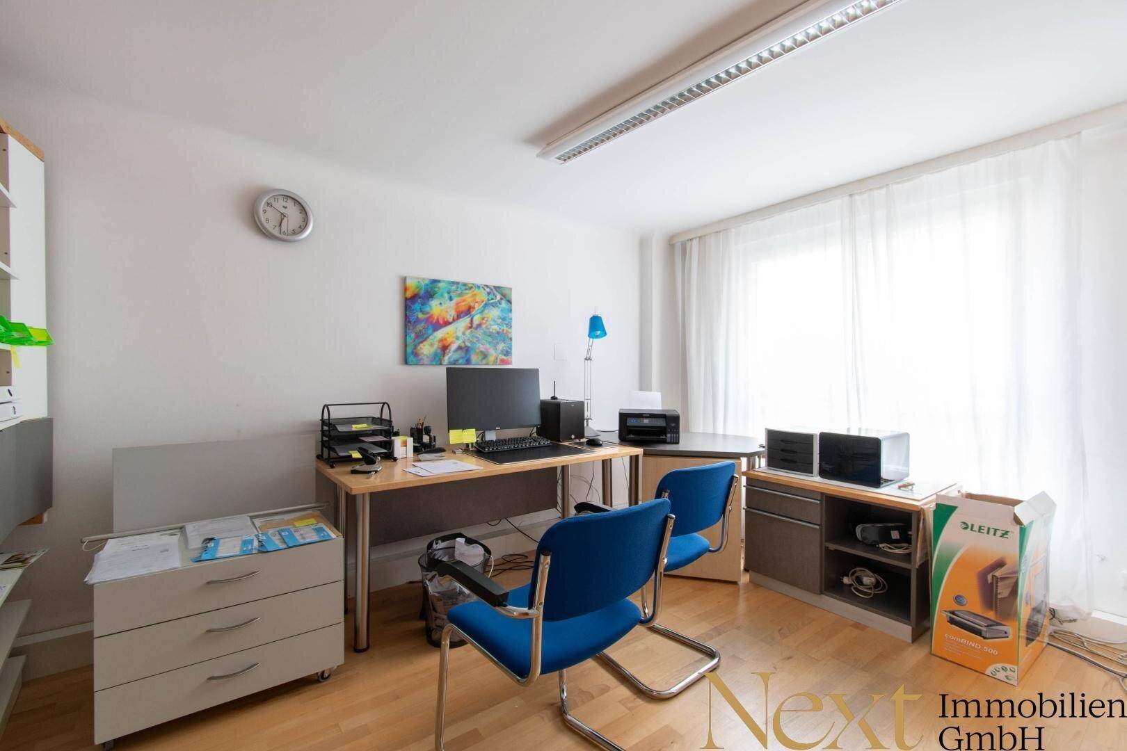 Helles Büro mit Holzfußboden, Schreibtisch, blauen Stühlen und großem Fenster mit weißen Vorhängen.