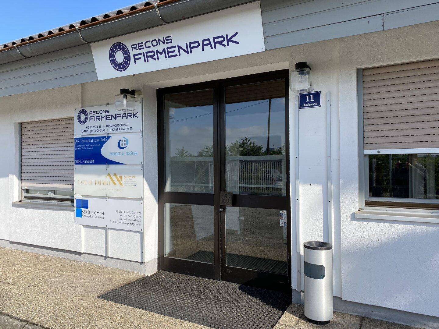 Eingangsbereich eines Firmenparks mit Glastüren, Beschilderung und Außenjalousien an den Fenstern.