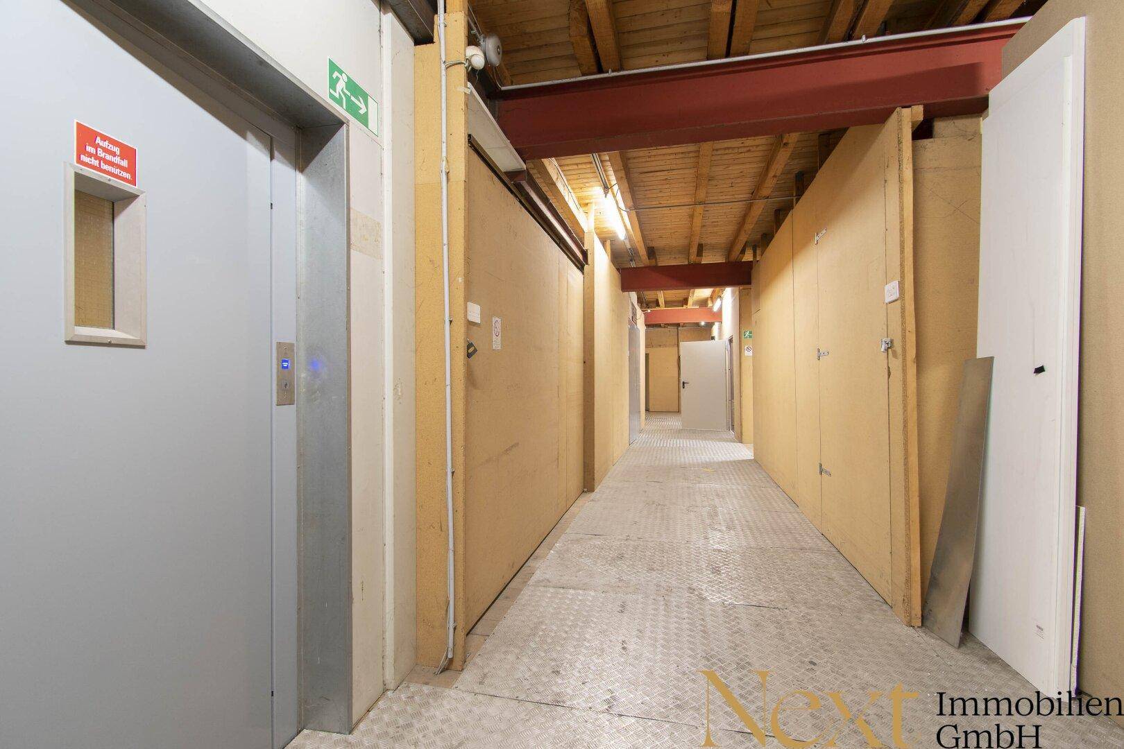 Langer Flur im Untergeschoss mit Aufzug, Lagerabteilen und sichtbaren Deckenbalken.