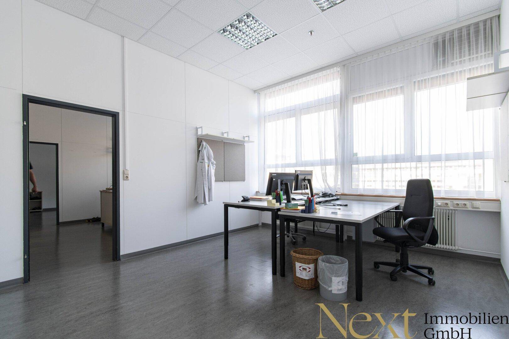 Helles Büro mit zwei Arbeitsplätzen, großen Fenstern und funktionaler Einrichtung.