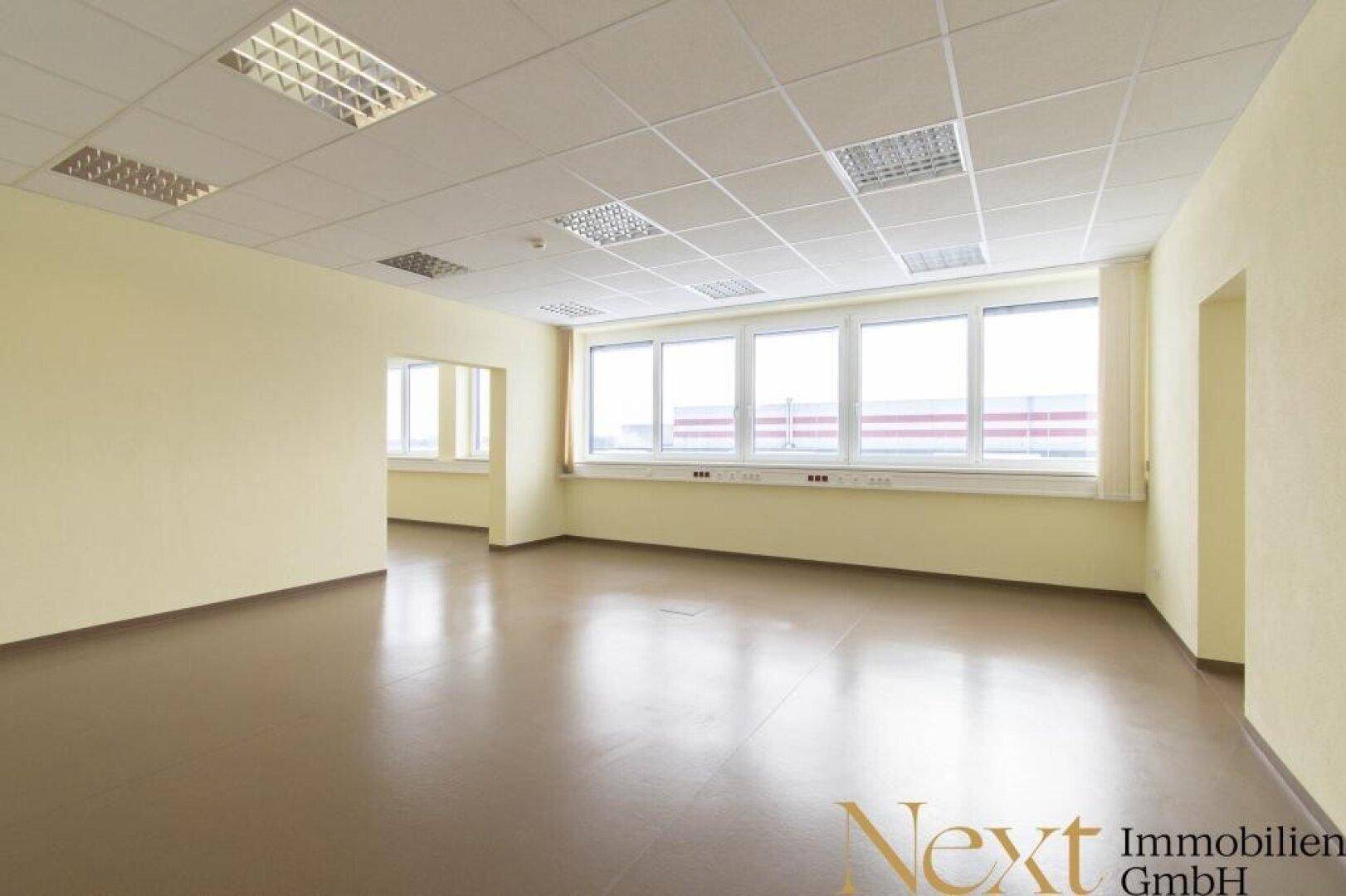 Geräumiges, leeres Büro mit großen Fenstern, die viel Tageslicht hereinlassen und einem hellbraunen Boden.