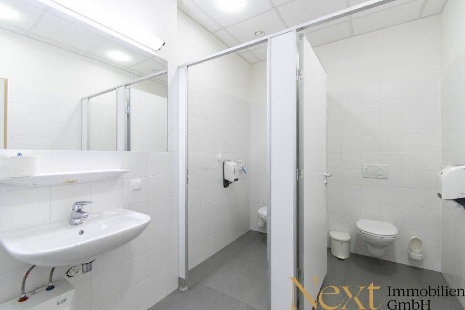Heller Sanitärbereich mit mehreren Toilettenkabinen, Waschbecken und weißen Fliesen.