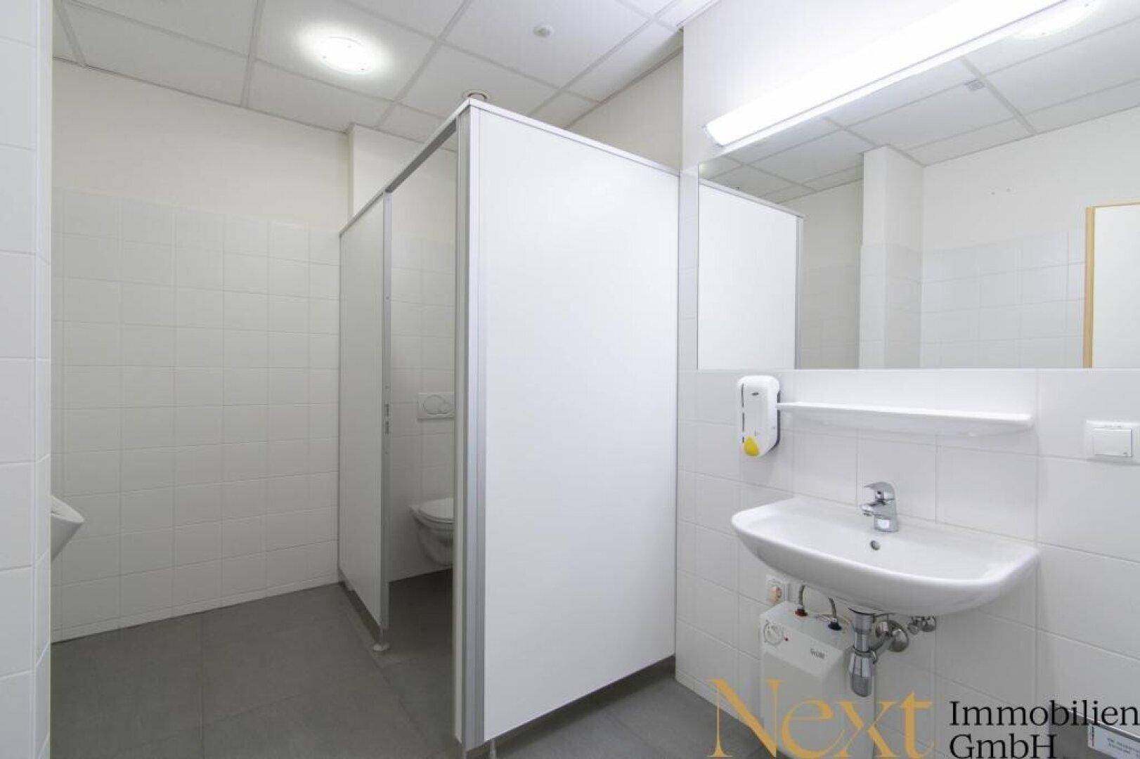Heller Sanitärbereich mit Toilettenkabinen, Waschbecken und weißen Fliesen.