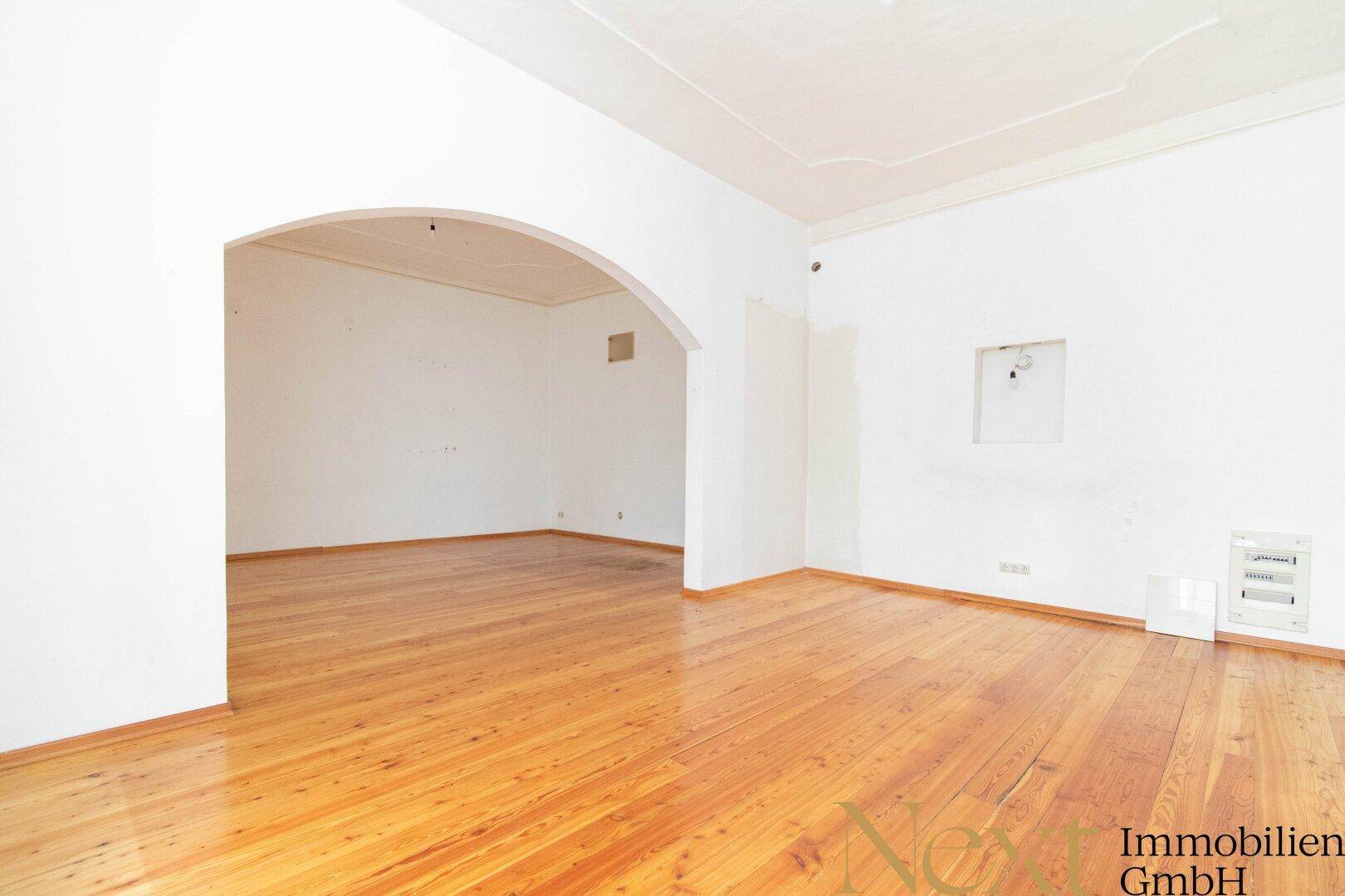 Heller Wohnraum mit Holzdielenboden und einem eleganten Bogen, der zu einem weiteren Bereich führt.