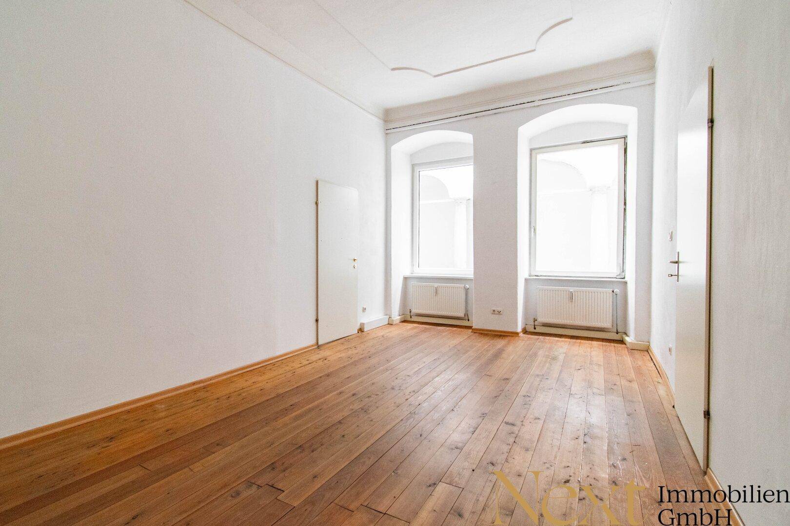 Geräumiger Raum mit Holzdielenboden, hohen Decken und zwei großen Bogenfenstern, die viel Licht spenden.