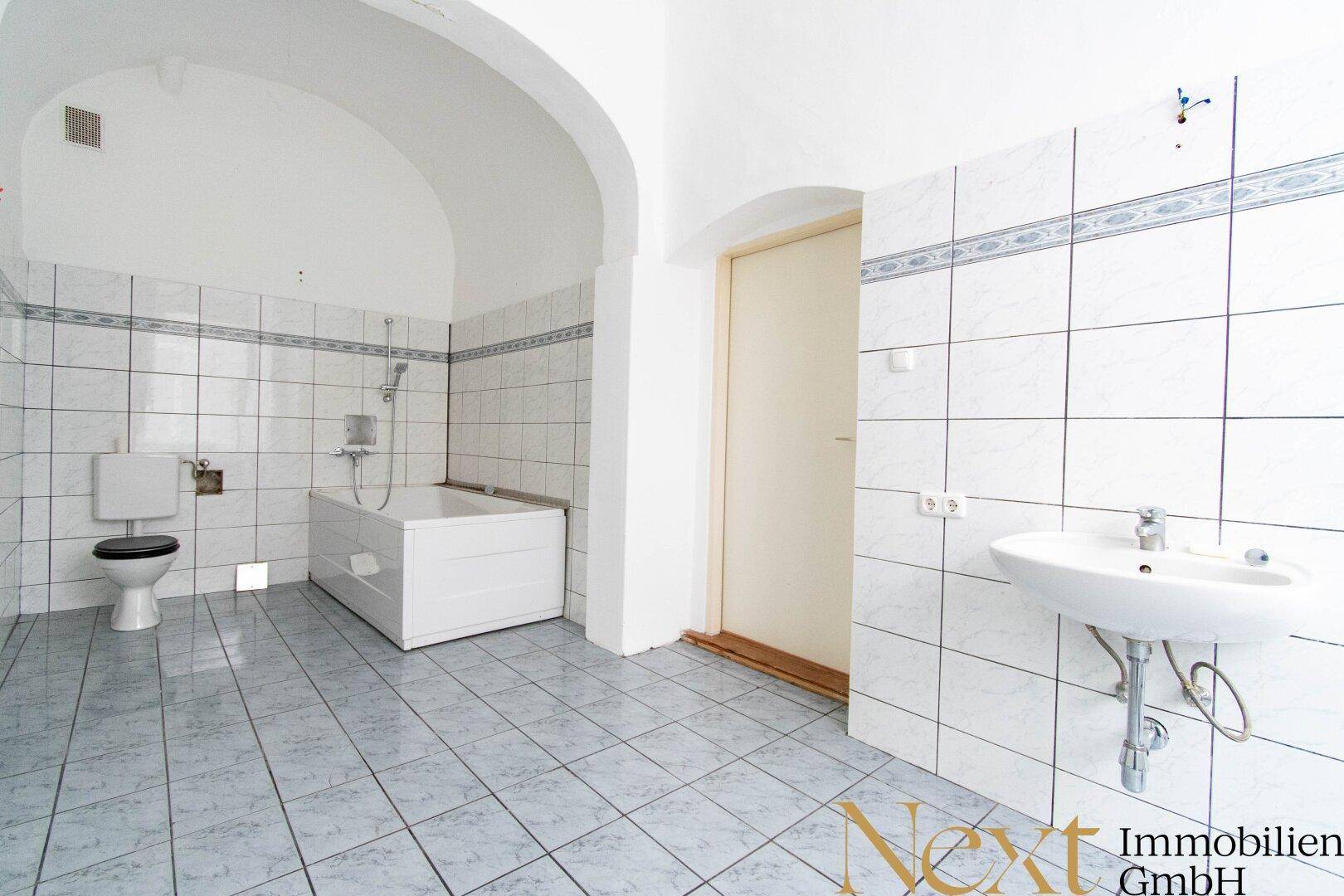 Gefliestes Badezimmer mit Badewanne, Waschbecken und Toilette unter einer Gewölbedecke, das renovierungsbedürftig ist.