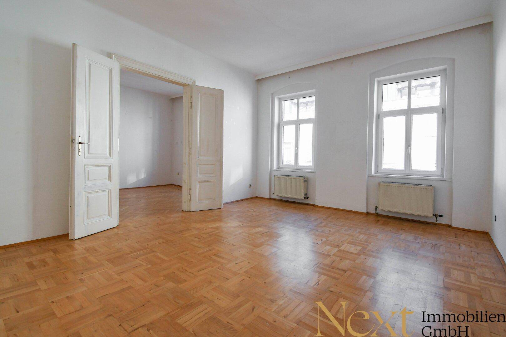 Heller Raum mit klassischem Parkettboden und hohen Doppeltüren, die zu einem weiteren Zimmer führen.